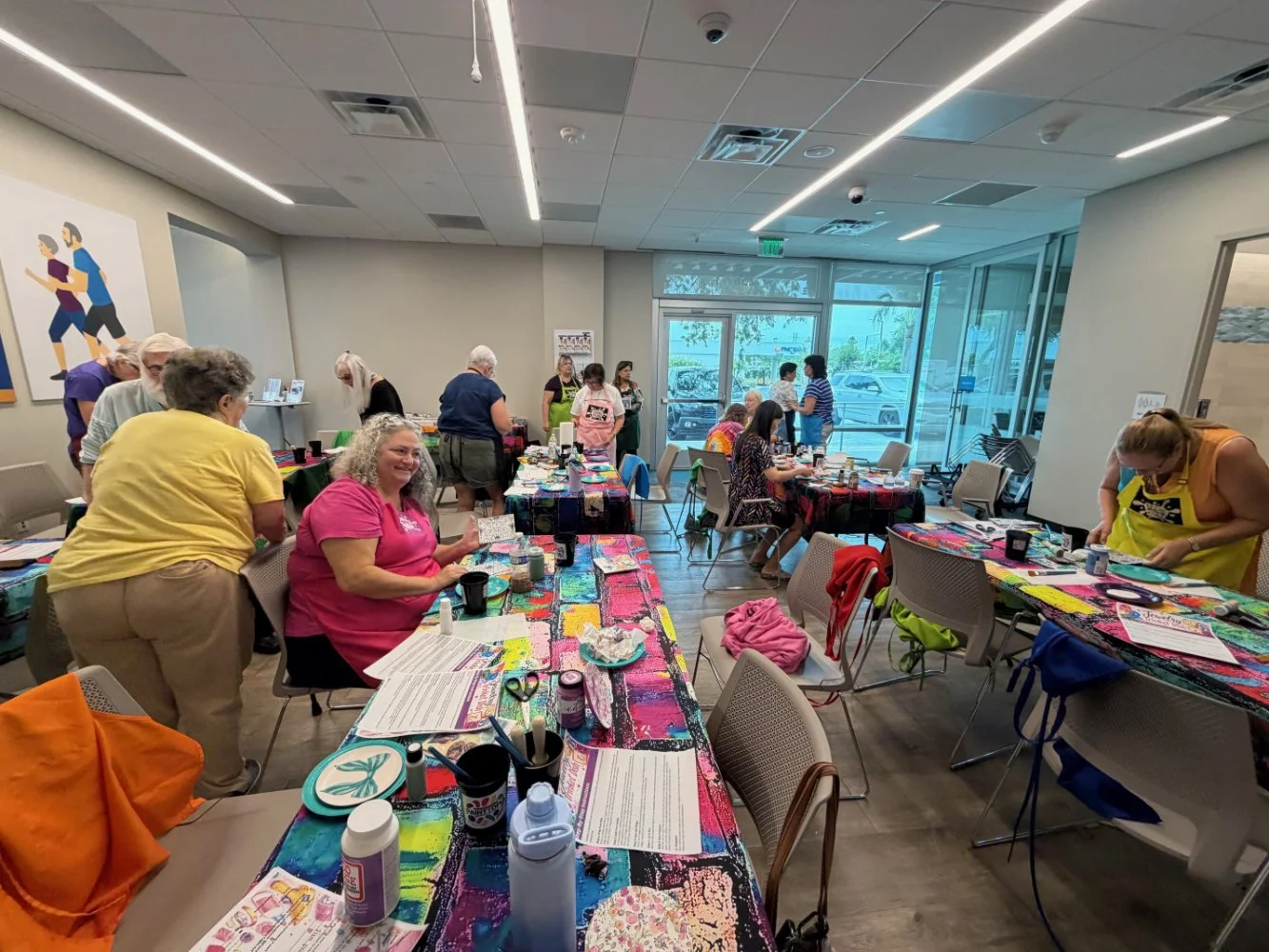 People gathered in a brightly lit indoor room with large windows, sitting and standing around tables covered with colorful tablecloths, engaging in arts and crafts activities.
