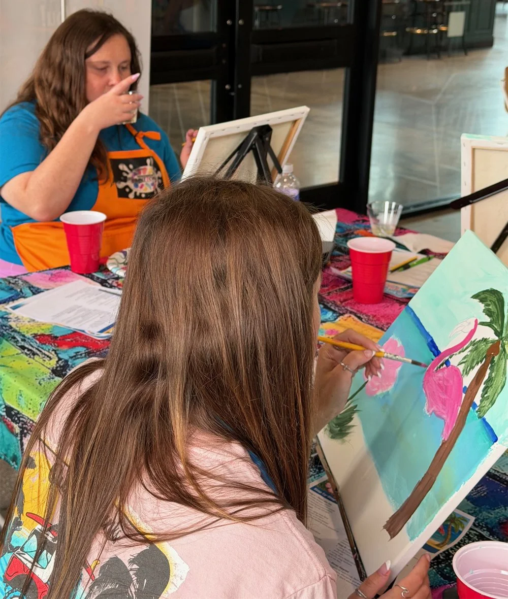 Two women painting tropical-themed pictures, one with a pink flamingo and the other with a colorful background, at a table covered with a colorful tablecloth, cups, and art supplies.