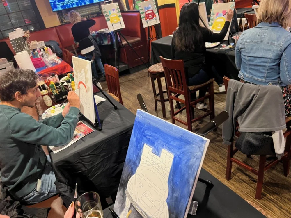 People participating in a painting class in a restaurant or bar, with canvases featuring car illustrations and colorful backgrounds, while drinks and art supplies are on the tables.