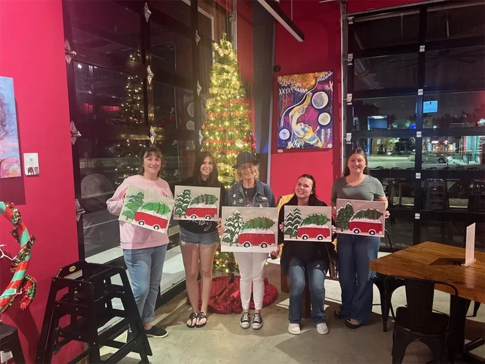 Group of adults and children holding colorful paintings of whimsical animals sitting on clouds, in a decorated room with tables and a sign that reads 'Boca Ciega' and 'Coquina Key'.