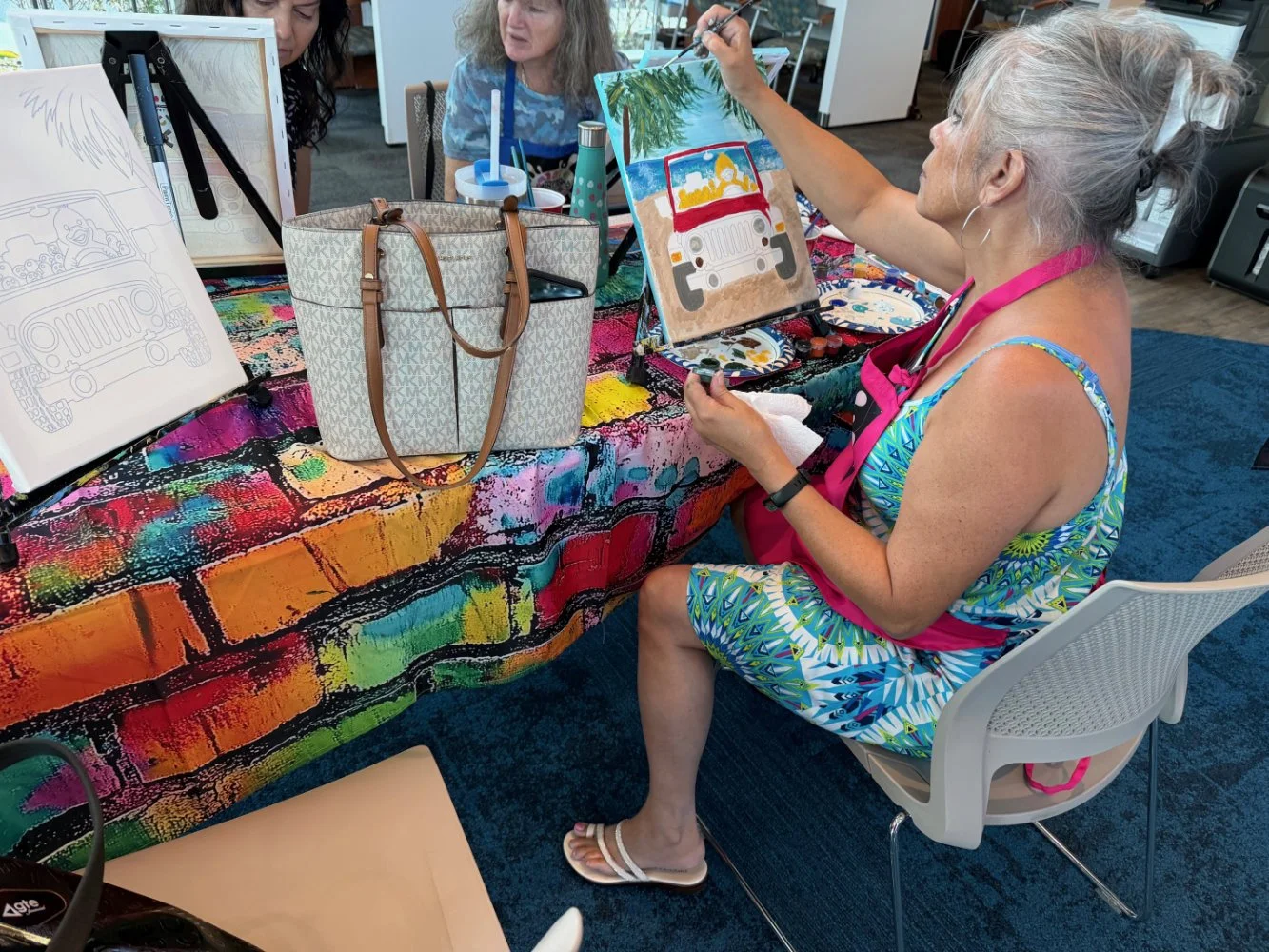 A woman with gray hair, wearing a colorful dress and a pink apron, is sitting at a table while painting a picture of a jeep on a canvas. She is holding a paintbrush and focusing on her artwork. There are two other women observing her, and the table i