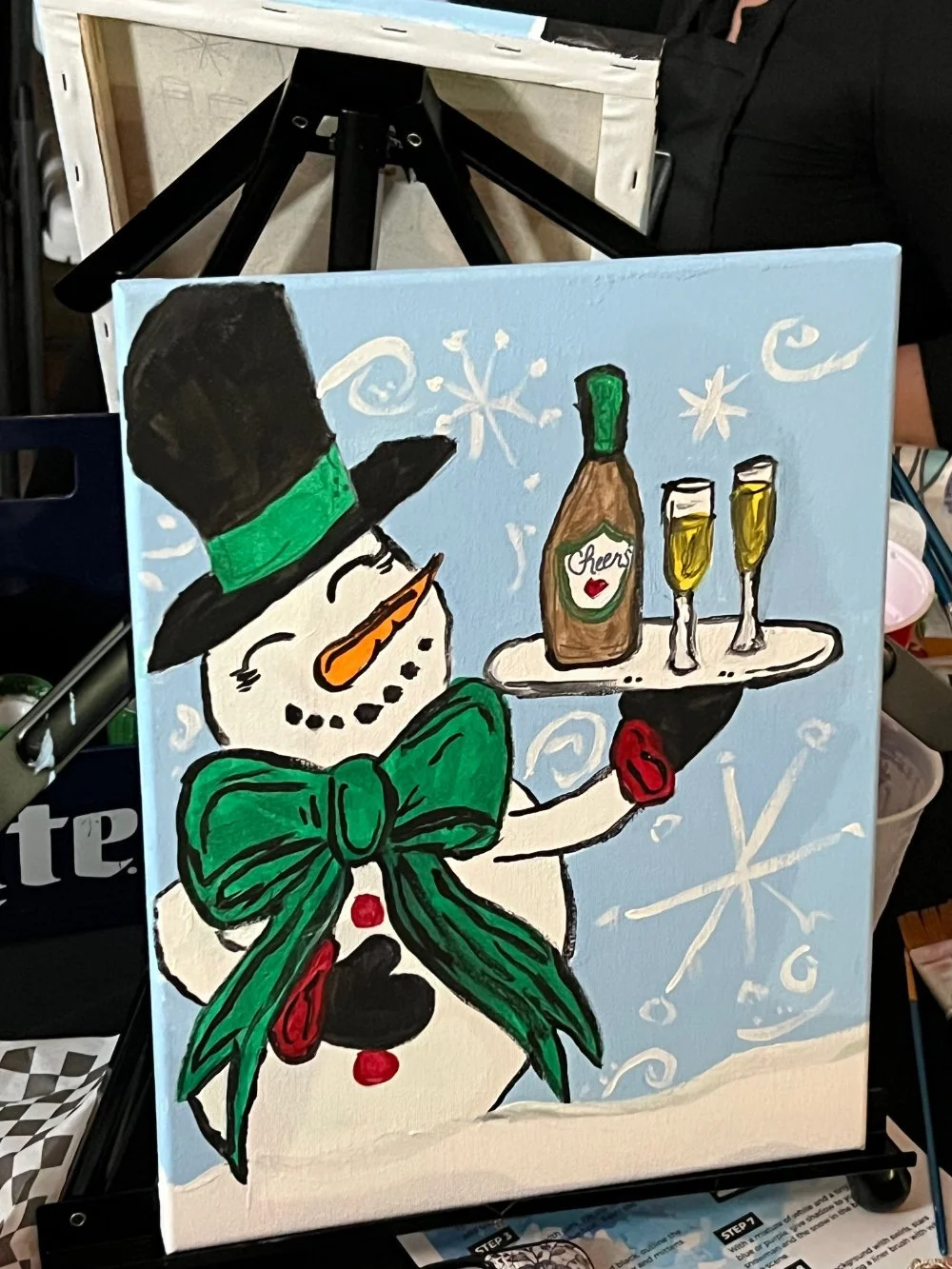 Painting of a snowman wearing a top hat with a green band, a large green bow tie, and red buttons. The snowman is holding a tray with a bottle labeled 'Cheers' and two glasses of champagne, with a snowy background and snowflake decorations.
