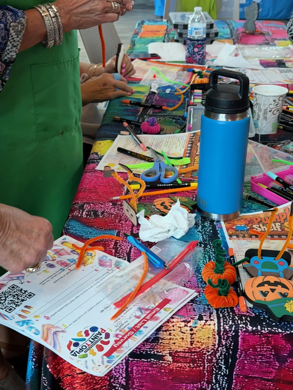 Table covered with arts and crafts supplies including pipe cleaners, markers, scissors, paper, and Halloween-themed decorations, with people working on craft projects.