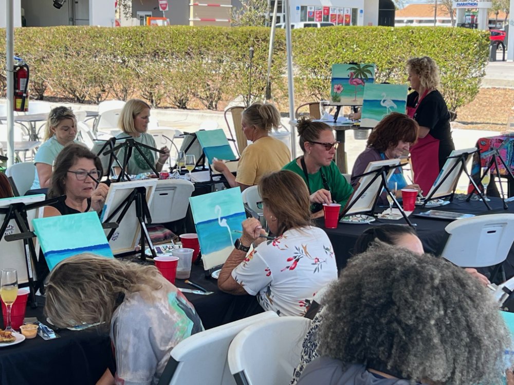 People painting at an outdoor art class, with paintings of beach scenes, palm trees, and flamingos visible.