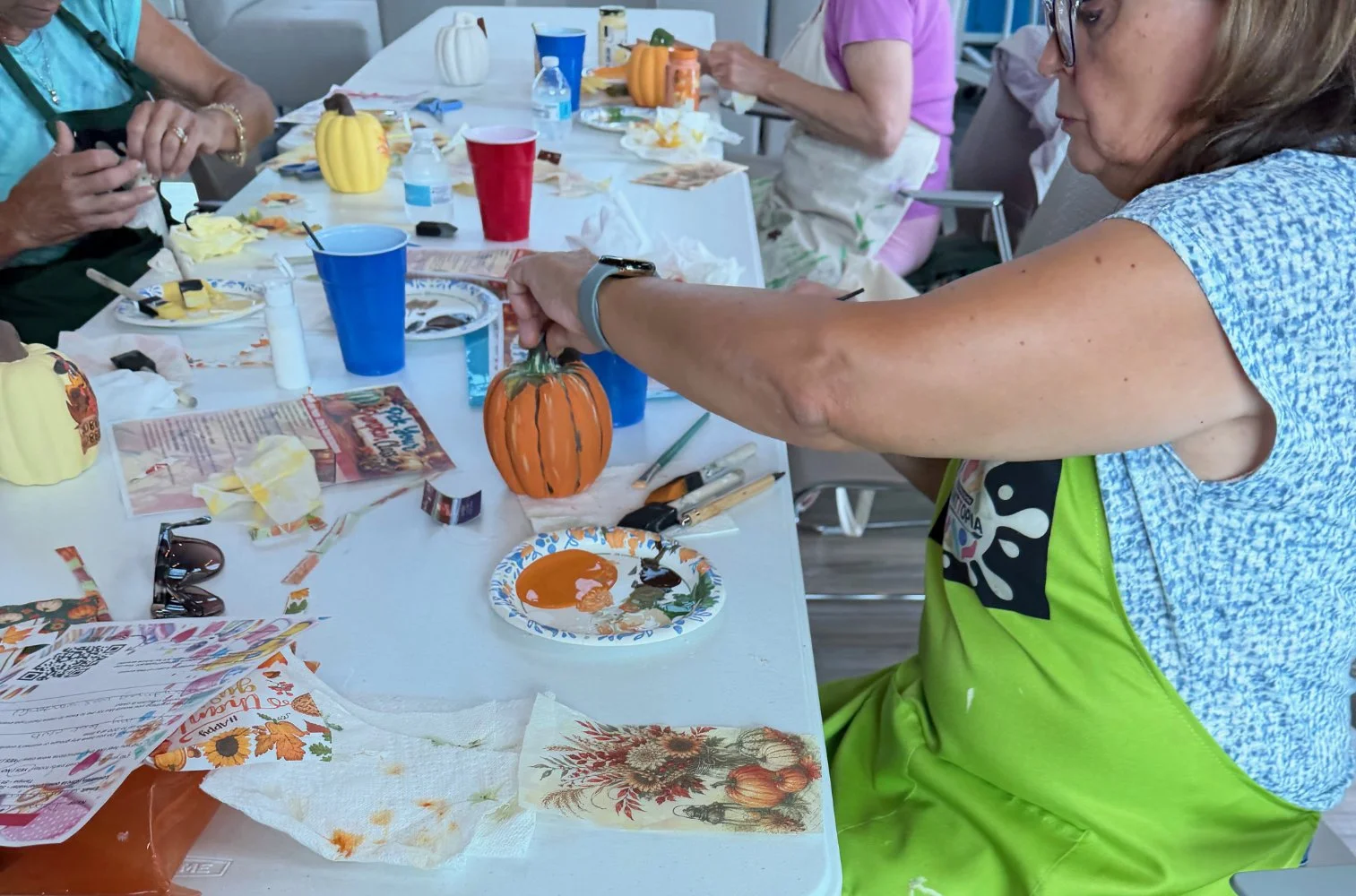 A woman painting or decorating a small pumpkin at a table with arts and crafts supplies, surrounded by other pumpkins and decorative items.