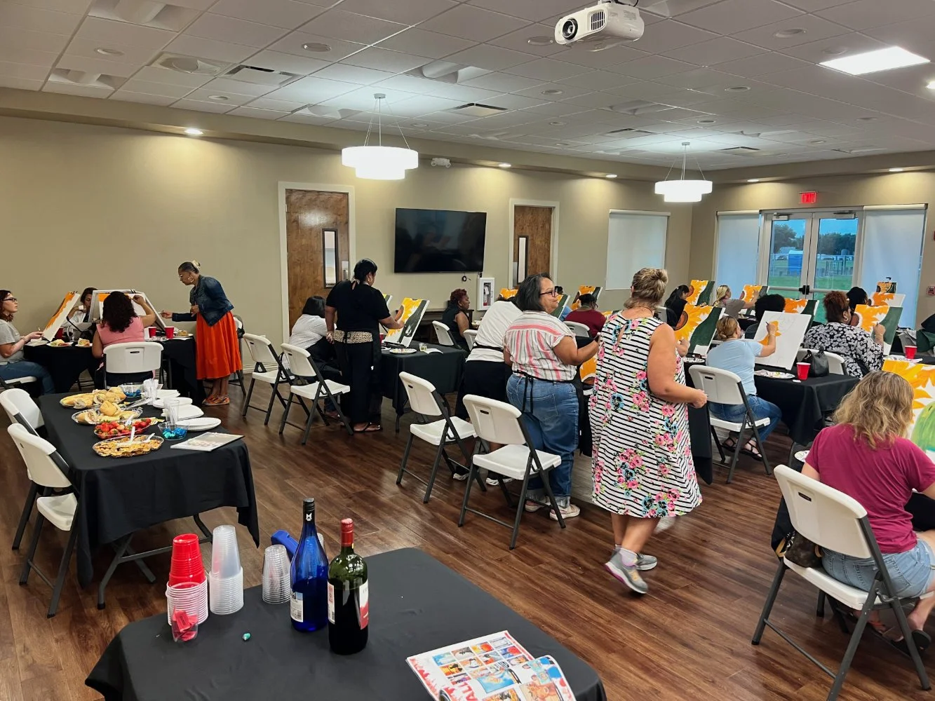 A group of people painting canvases at tables in a spacious, well-lit indoor room with large windows. Some tables have snacks and drinks, and a TV is mounted on the wall.