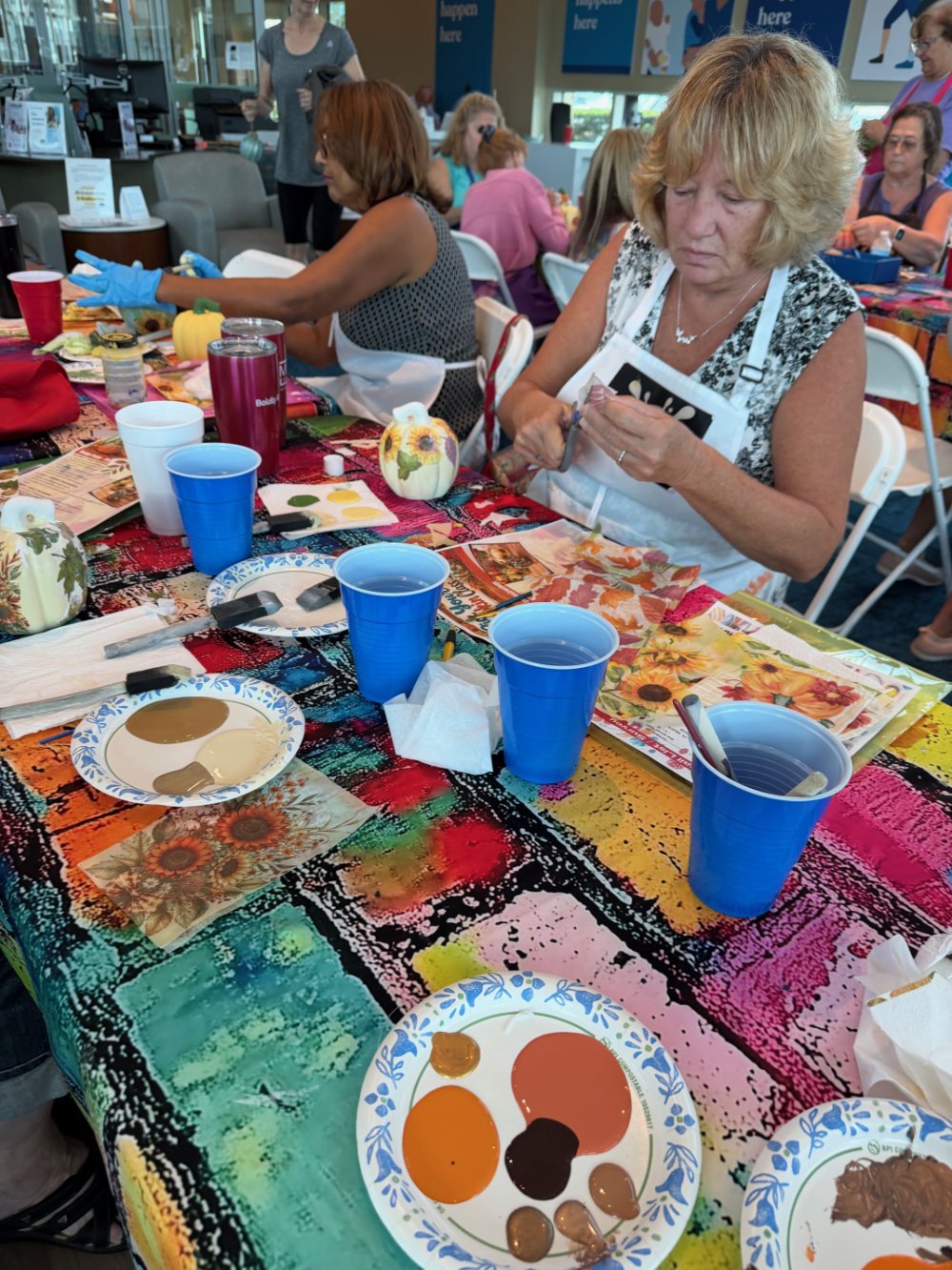 People participating in a fall craft event at a colorful table with paint, brushes, and paper, in a bright indoor setting.