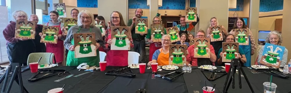 Group of people at a Christmas-themed party holding paintings of a Christmas elf with a green hat and collar, smiling and posing for the camera in a decorated room.
