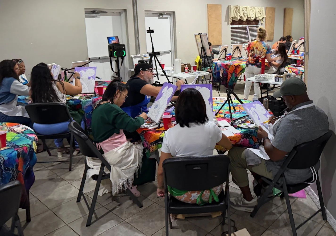 People participating in a group painting class, sitting at tables covered with colorful tablecloths, working on paintings of purple unicorns on canvases in a well-lit room with window and bulletin board.