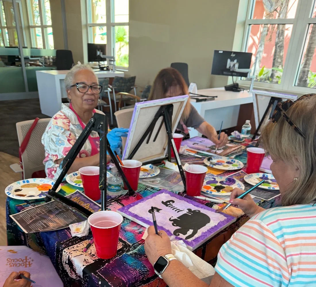 Group of women painting silhouettes of elephants on canvases at a table during a painting class.