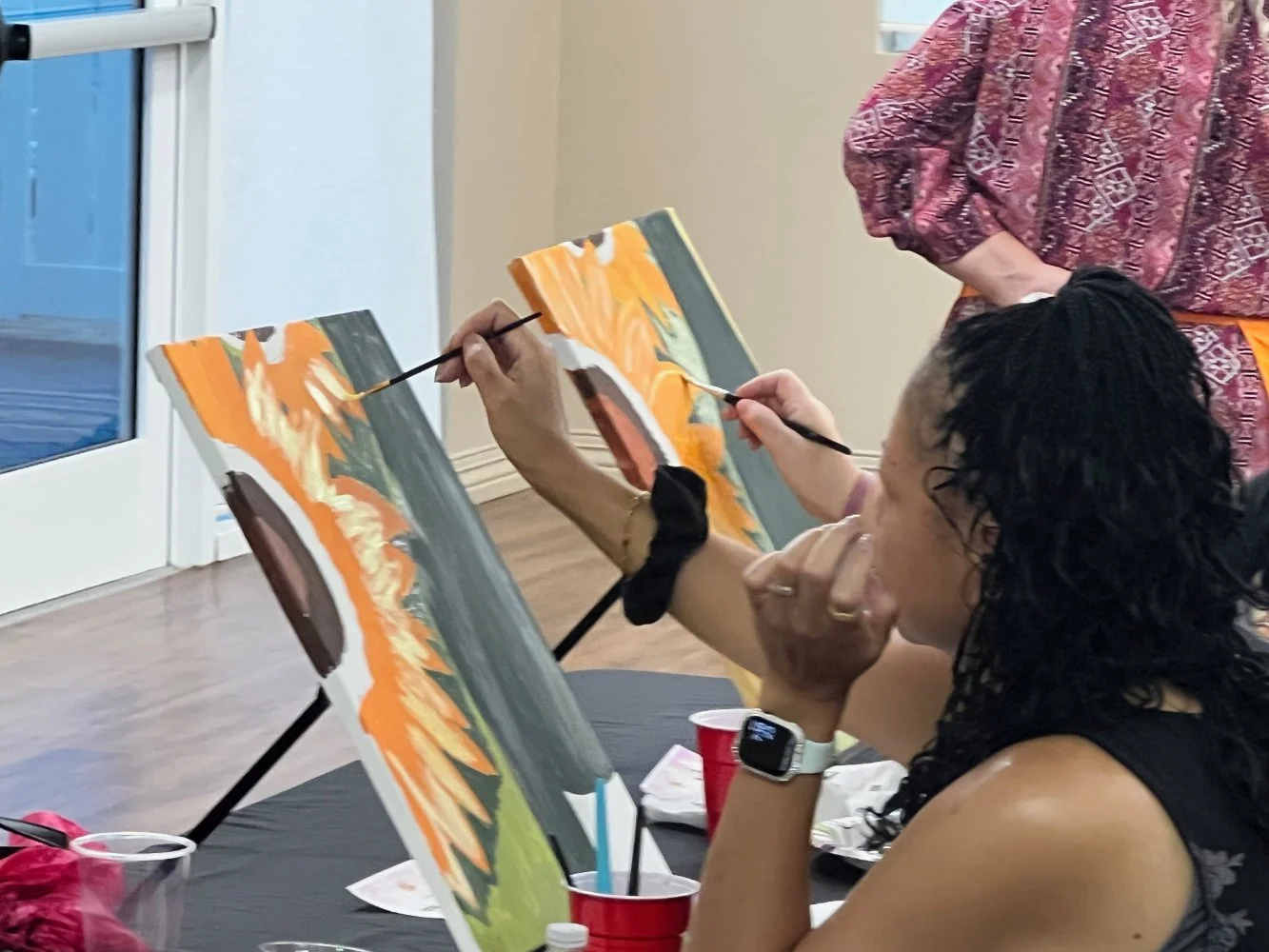 A woman with curly dark hair is painting on a canvas at a painting party, with colorful abstract designs. She is wearing a wristwatch and a sleeveless top, with her hand resting on her chin, focused on her artwork. Another person stands nearby giving