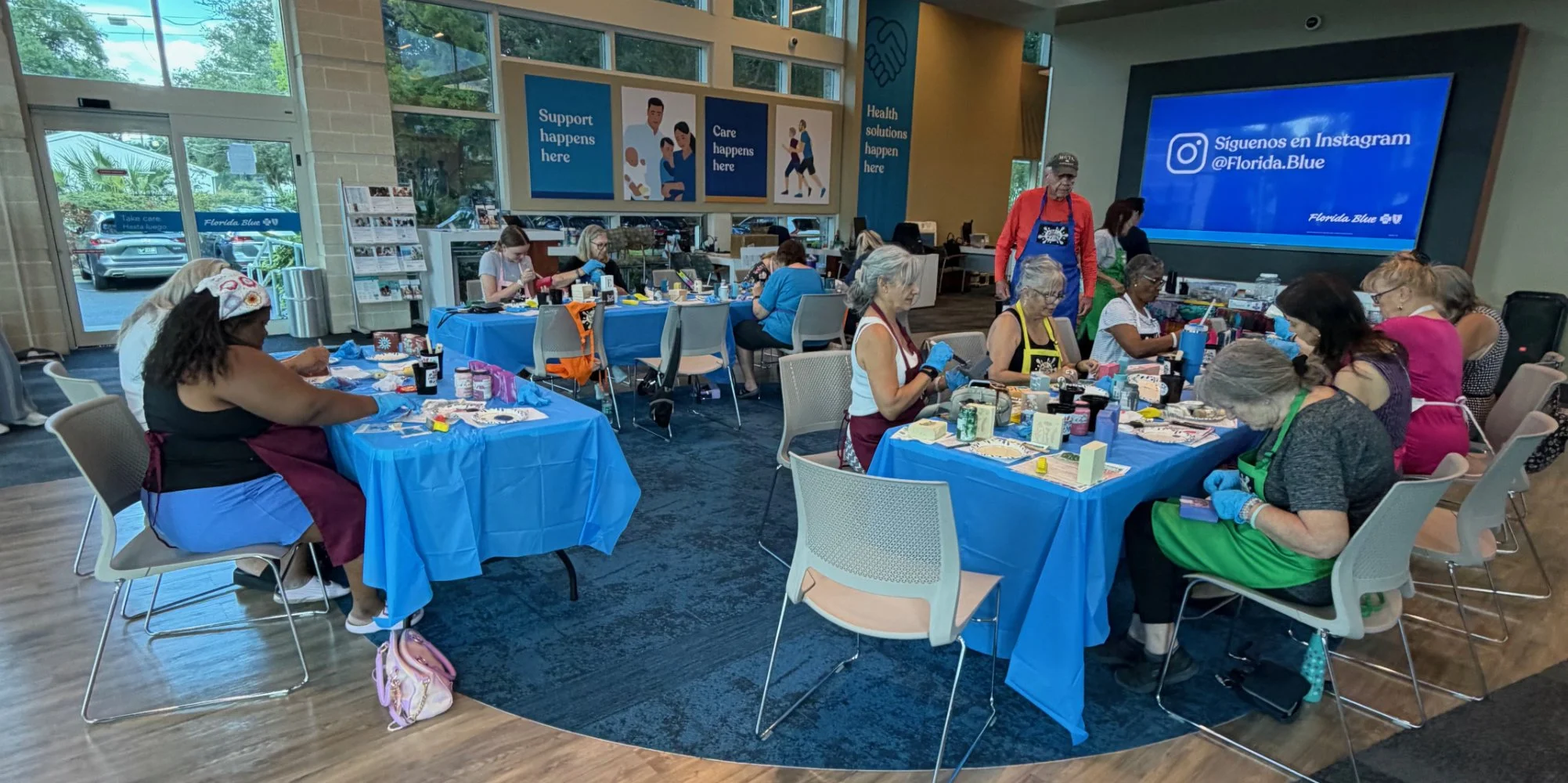 Group of women participating in a crafting activity at tables covered with blue tablecloths inside a spacious room with large windows. The room features various signs and a large screen displaying Instagram handle for Florida Blue.