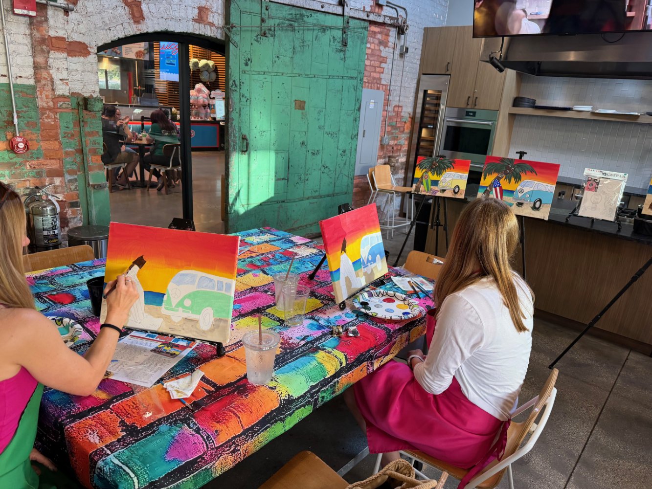 Two women are sitting at a table in an art studio, painting picture of a colorful camper van with sunset backgrounds on small canvas boards. The table is covered with a vibrant, multicolored tablecloth, and painting supplies are visible. Other painti