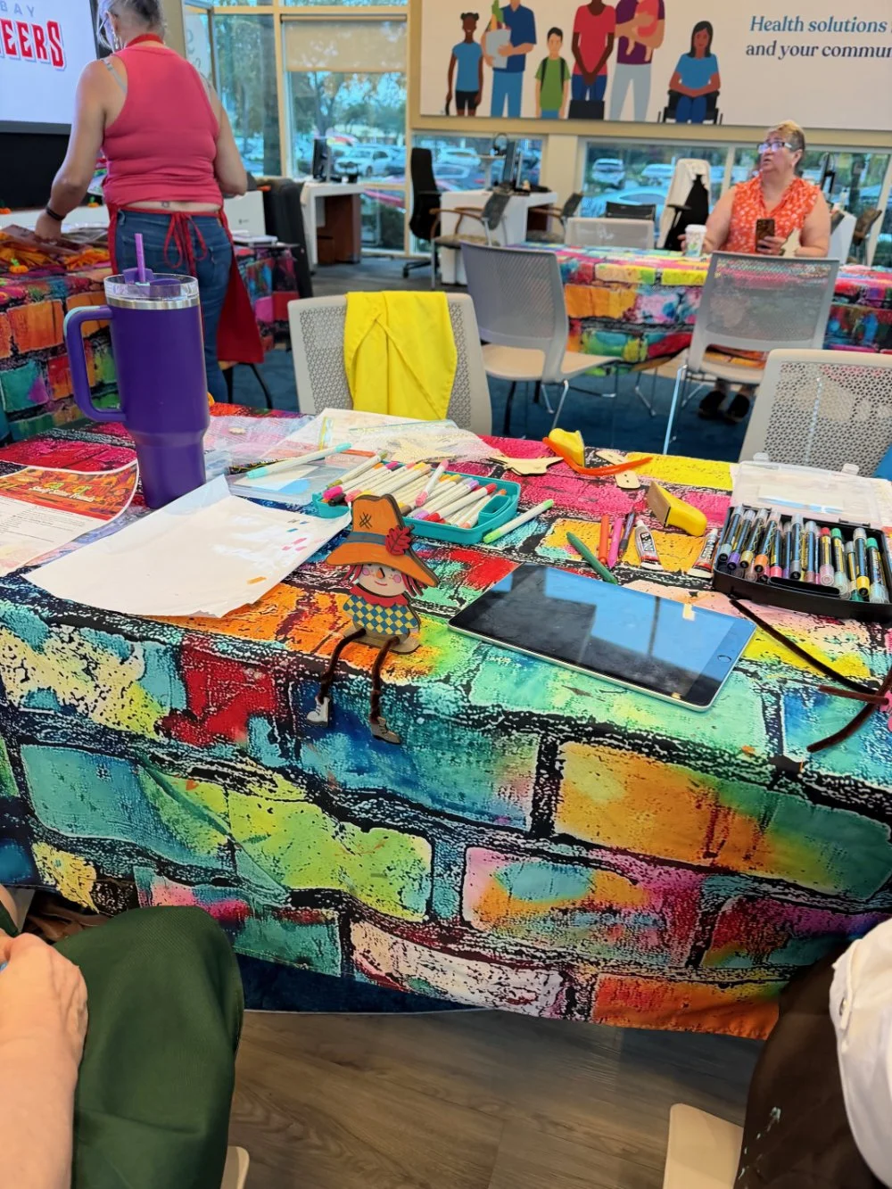 A colorful table with art supplies including markers, paper, and a tablet, in a room with people and a large mural of diverse people in the background.