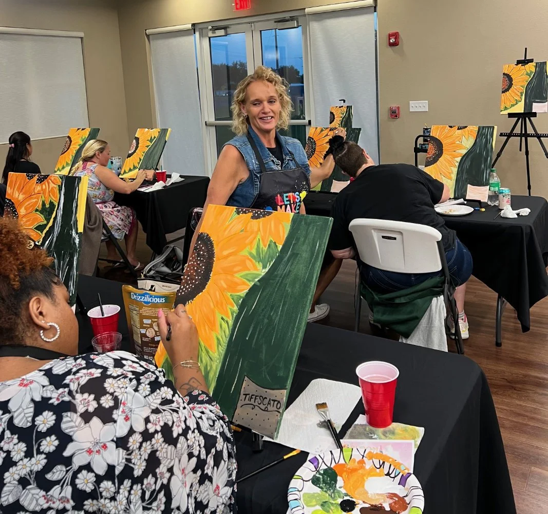 Women participating in a painting class, painting sunflower pictures on canvases, with art supplies and drinks on tables in a well-lit room.