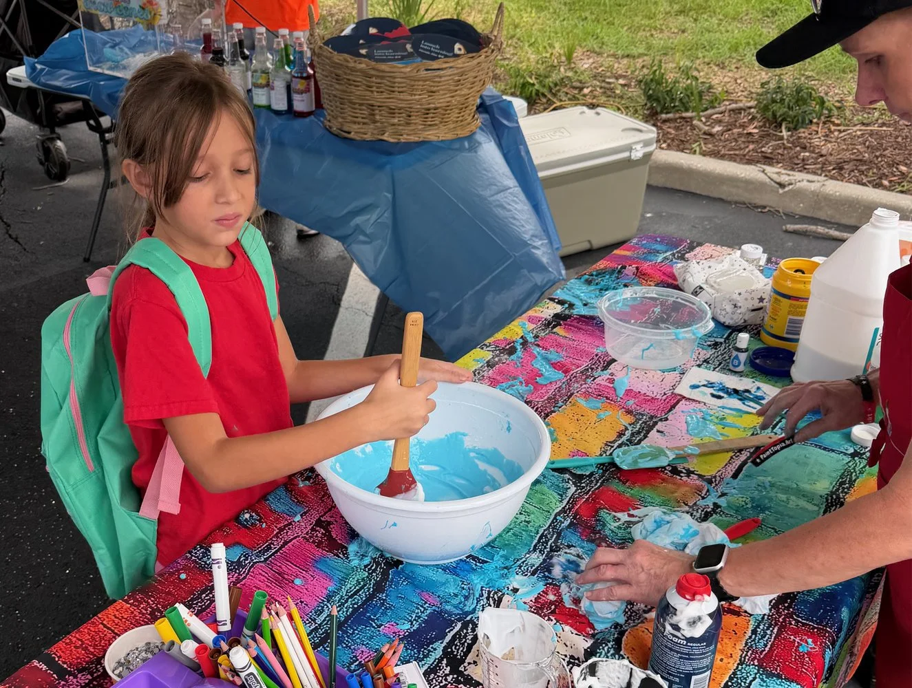 A young girl with a green backpack standing next to a table covered with colorful paint and art supplies, mixing blue paint in a white bowl, while an adult nearby is also working with paints outside.