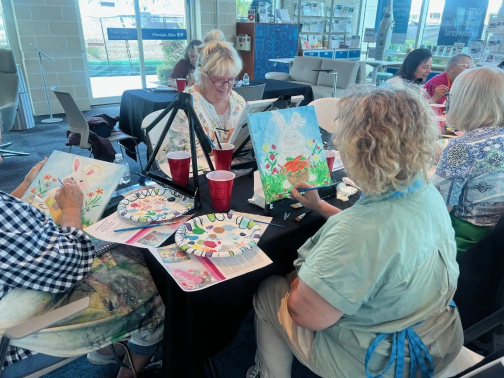 Group of people participating in a painting class, creating colorful artwork in a well-lit indoor space with large glass windows.