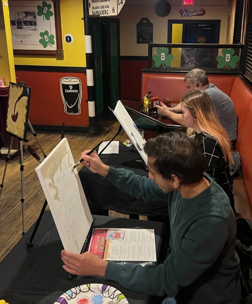 People painting festive holiday canvases at a restaurant or pub with beer and condiment bottles on the table, decorated with shamrocks and St. Patrick's Day theme.
