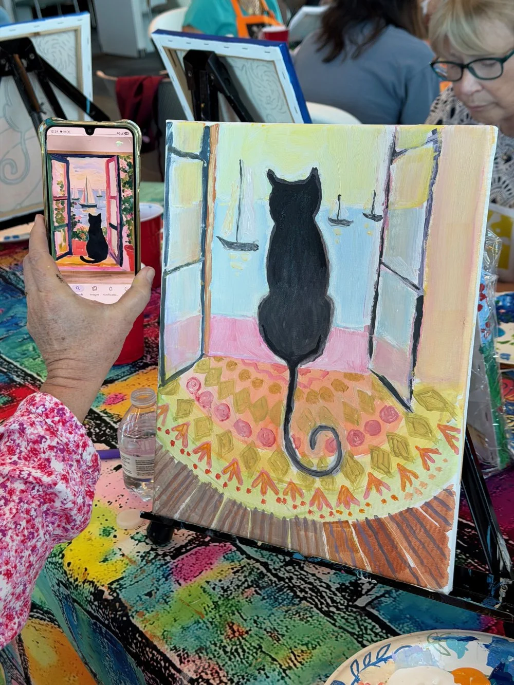 A person is taking a photo of a painting with a smartphone. The painting depicts a black cat sitting on a ledge, looking out an open window at sailboats on the water under a colorful sky. The scene is indoors, in an art class or workshop, with other 