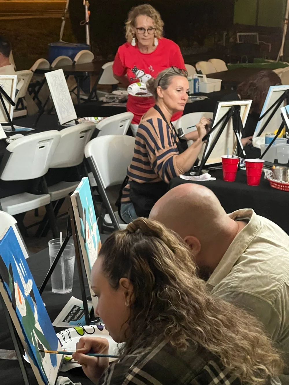 People participating in a painting class, painting on canvases at tables with art supplies, some with drinks, in an indoor setting.