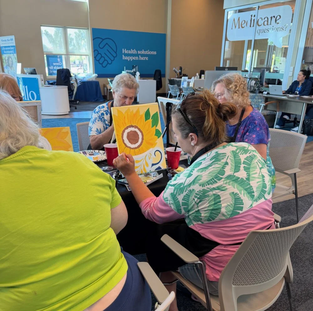 A group of people painting sunflower pictures at a table in a community center or office space with signs about healthcare in the background.