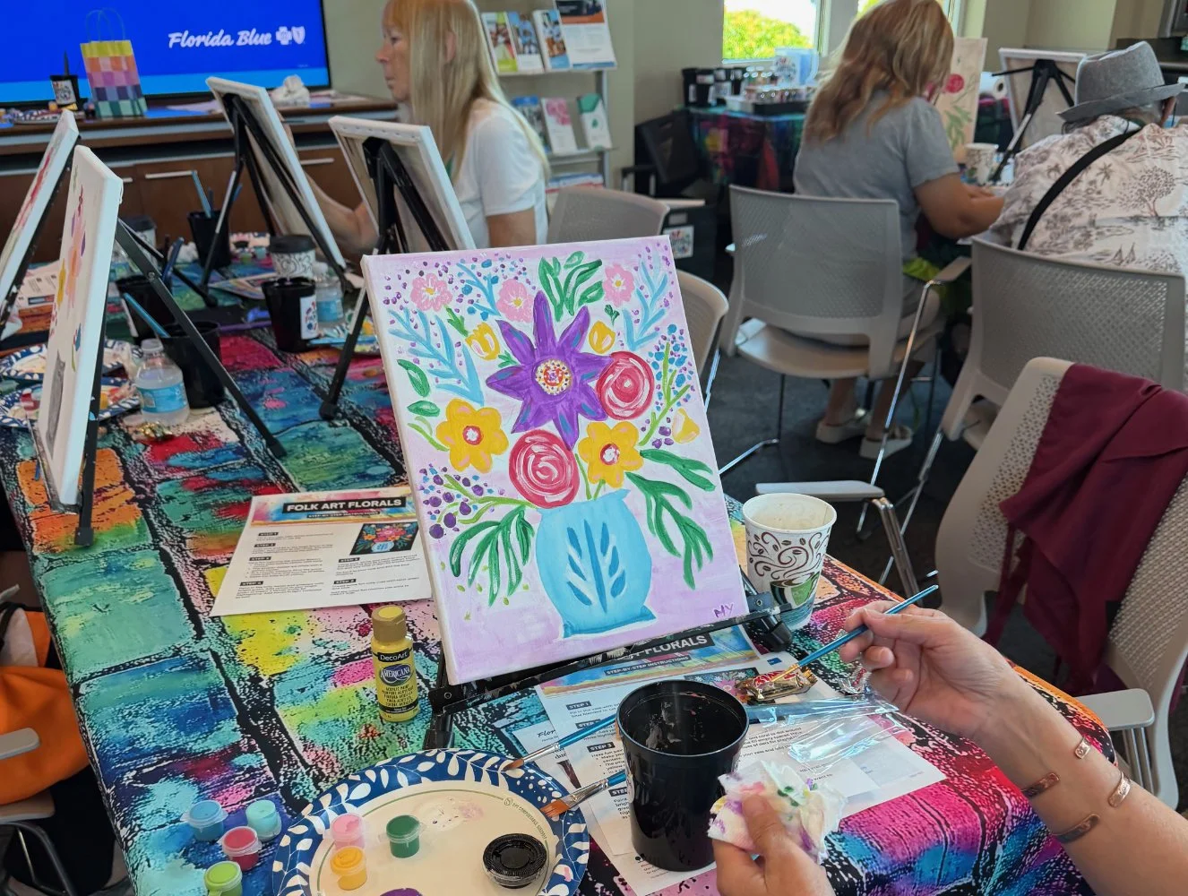 A colorful floral painting on an easel featuring purple, red, yellow, and pink flowers in a blue vase. The scene is set at a painting class with supplies on a vibrant, multicolored table. Several people are seated around the table, working on their o