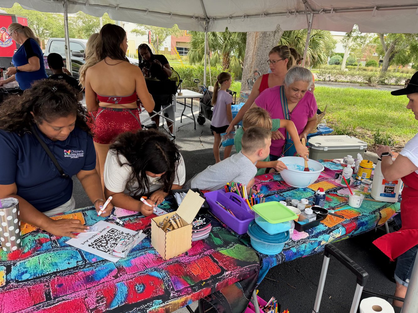 People gathered under a tent participating in a craft activity, including children and adults, with art supplies and colorful decorations on the table.