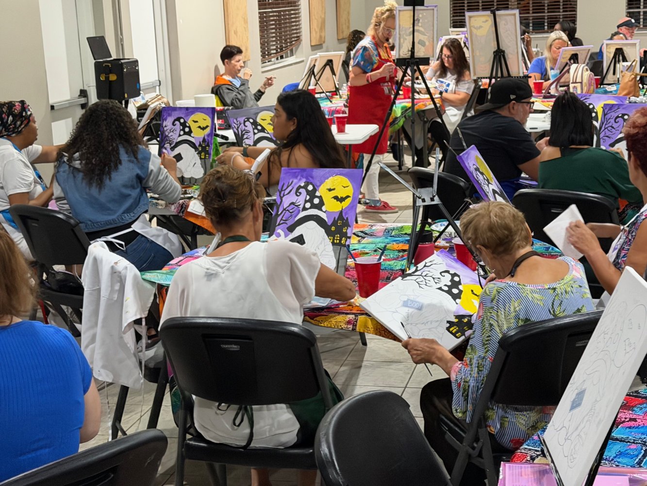 Group of people participating in a painting class, working on Halloween-themed canvases with a ghost, haunted house, bats, and a full moon.