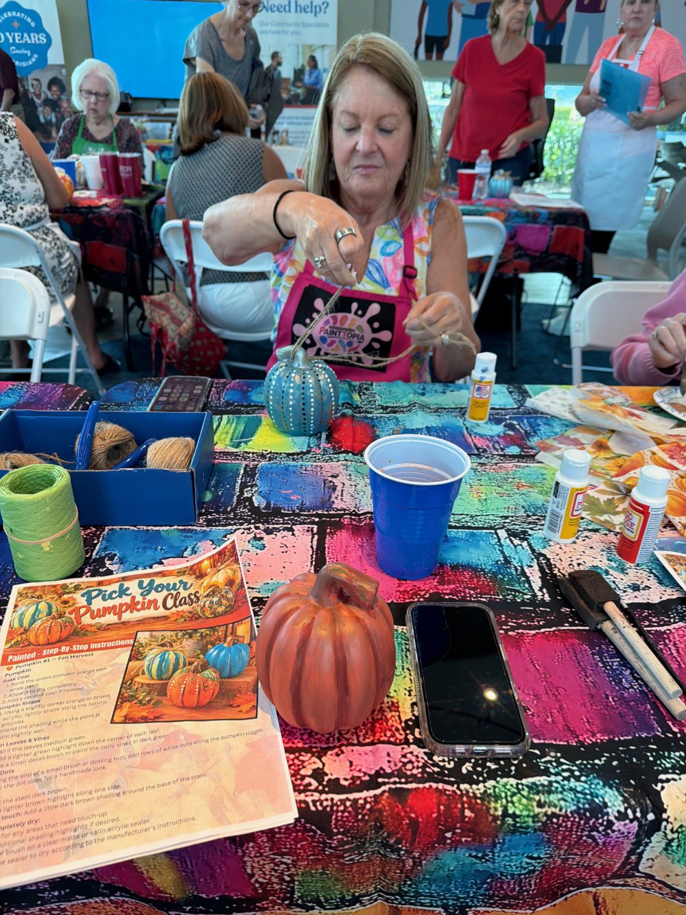 A woman participating in a painting class at a pumpkin-themed event, surrounded by art supplies and a colorful tablecloth with other people in the background.