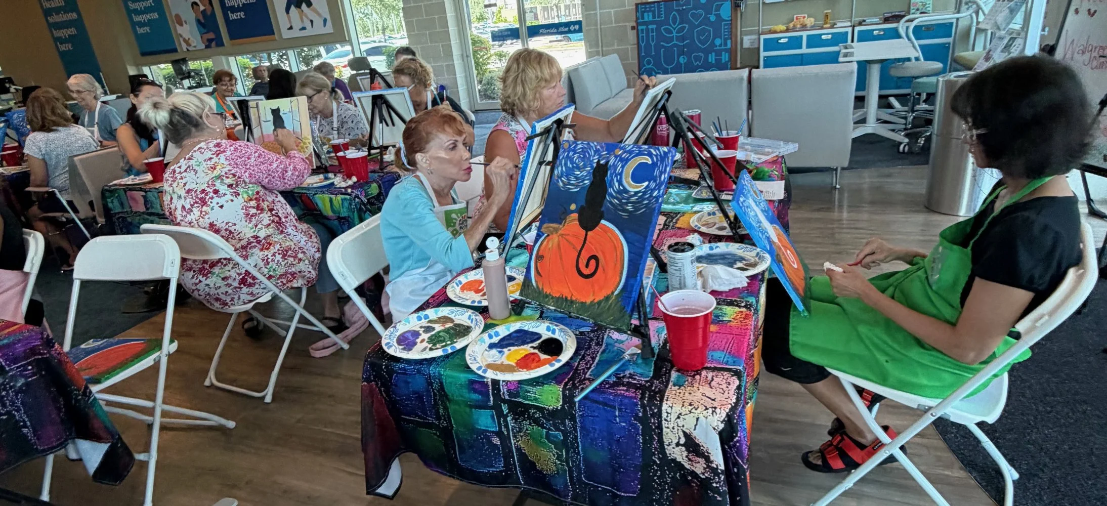 A group of women participating in a painting class indoors, with paintings of Halloween themes, including a pumpkin, a cat, and a moon, displayed on their canvases.