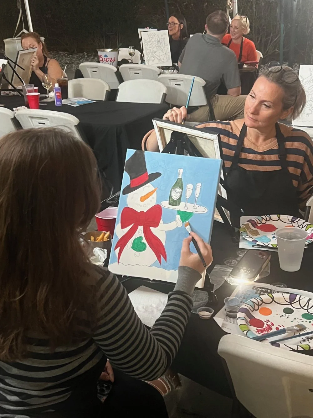 Woman painting a colorful snowman with a black hat, red bow, and green heart on a canvas at an art party.