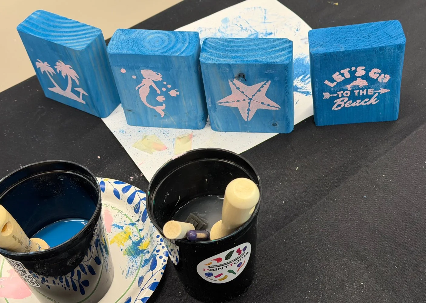 Decorative wooden blocks painted blue with white beach-themed designs, including palm trees, a mermaid, a starfish, and the phrase "Let's go to the beach." Two black paint cans with paint-stained lids and foam paintbrushes are in the foreground. The 