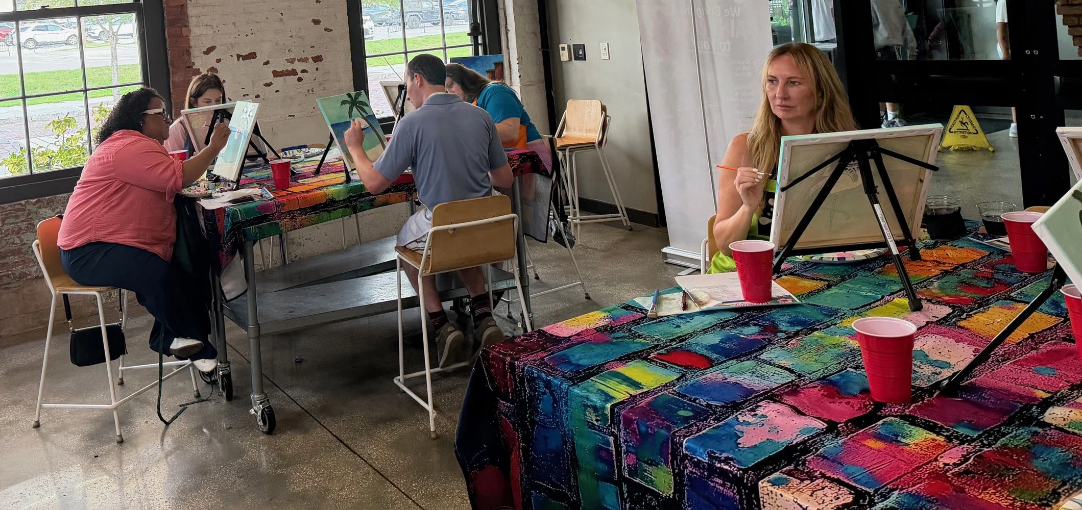 People painting on canvases at a colorful table in an art studio with large windows and exposed brick walls.