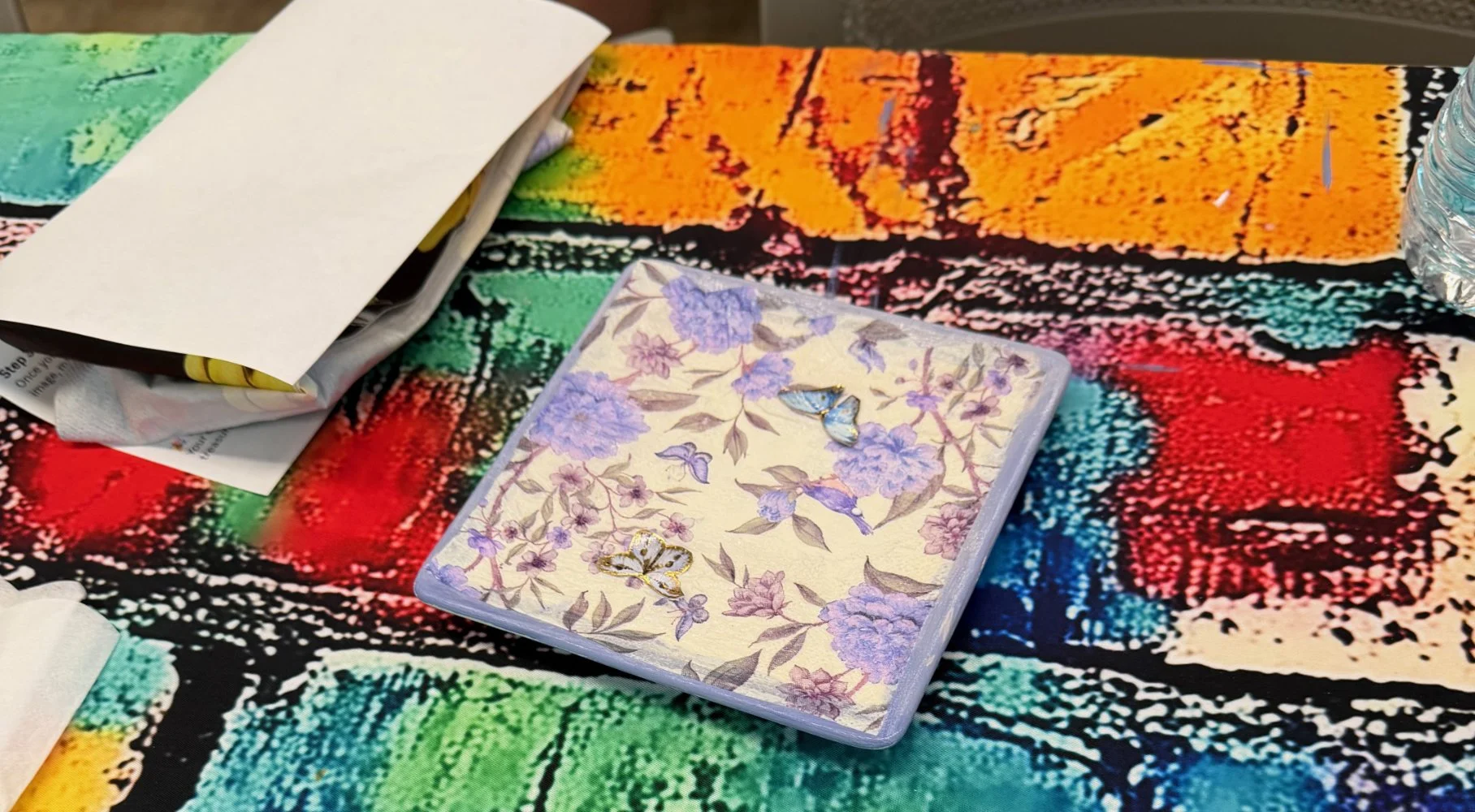 A table with a colorful, abstract patterned tablecloth, a white notebook or planner, a floral-patterned playing card or coaster featuring a butterfly and purple flowers, and a clear plastic water bottle.