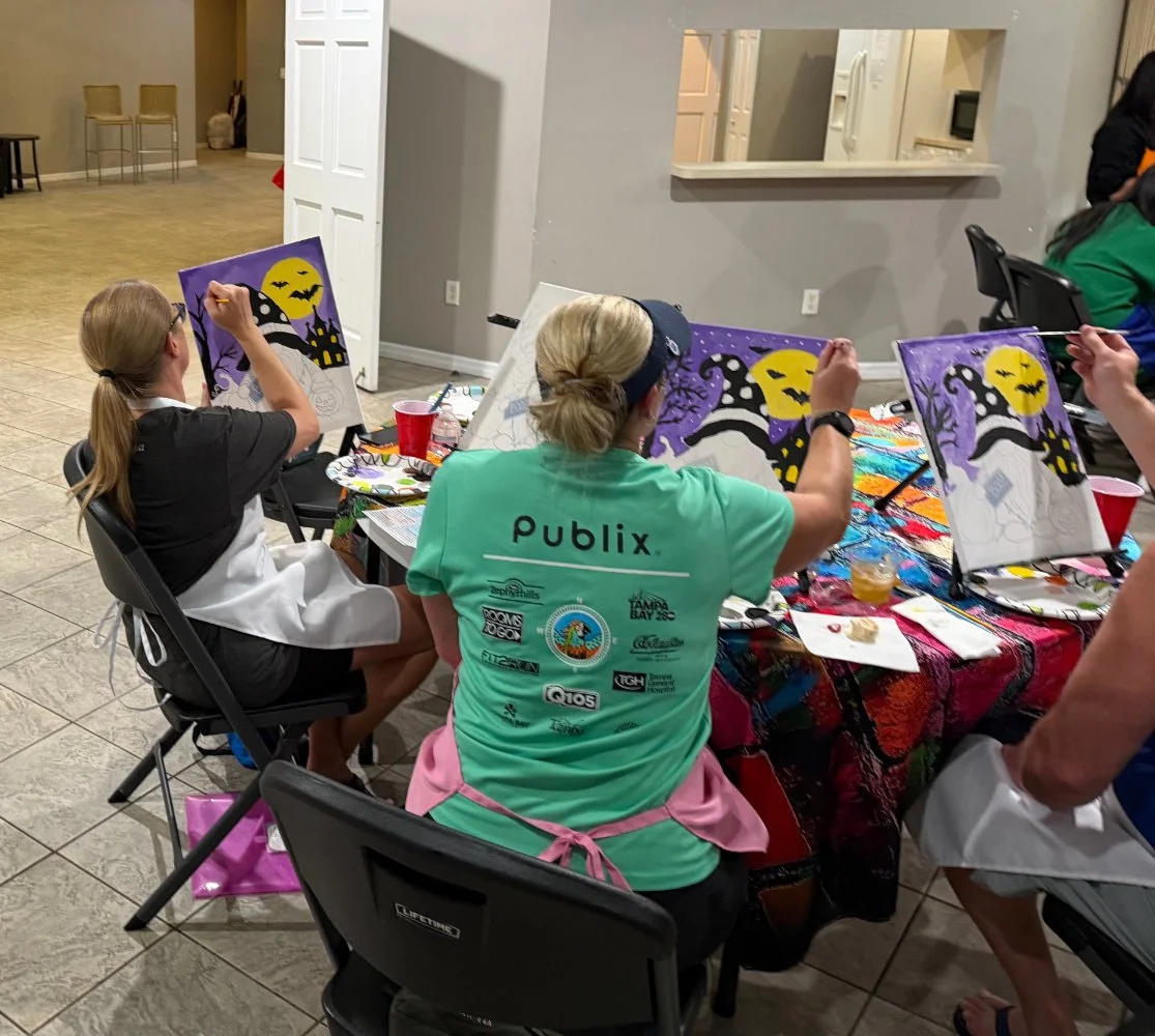 People painting Halloween-themed pictures with a witch silhouette, a full moon, and bats on canvases, seated around a colorful table in an indoor setting.