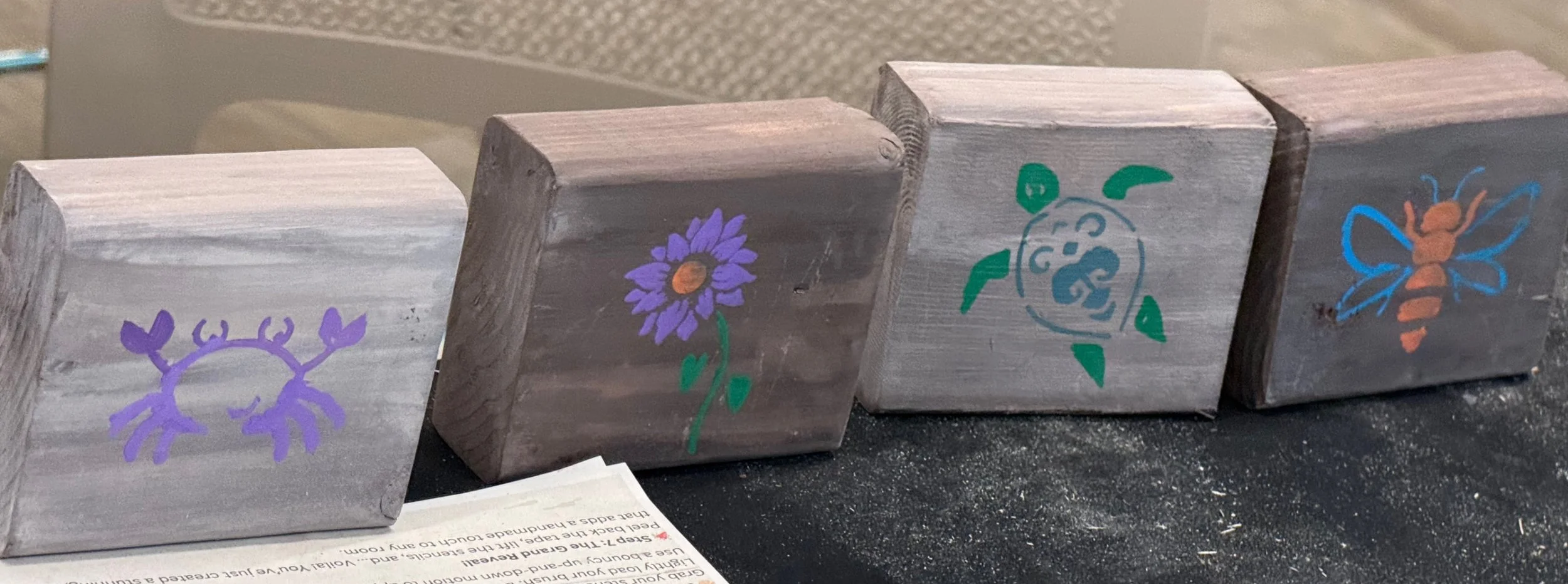 Four wooden blocks with hand-painted purple crab, purple flower, green snail, and orange bee illustrations.