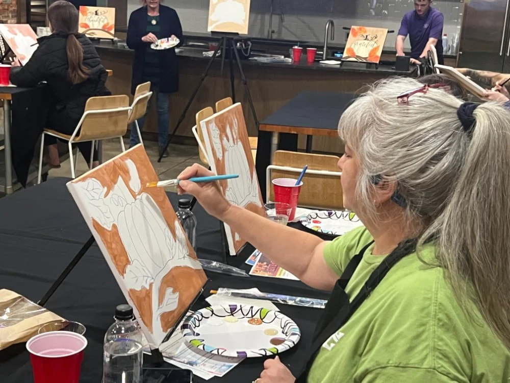 Woman with gray hair in a ponytail, wearing a green shirt and black apron, painting a pumpkin on canvas at a party. Other attendees are sitting at tables, and a woman in dark clothing stands in the background. The setting appears to be a casual art c