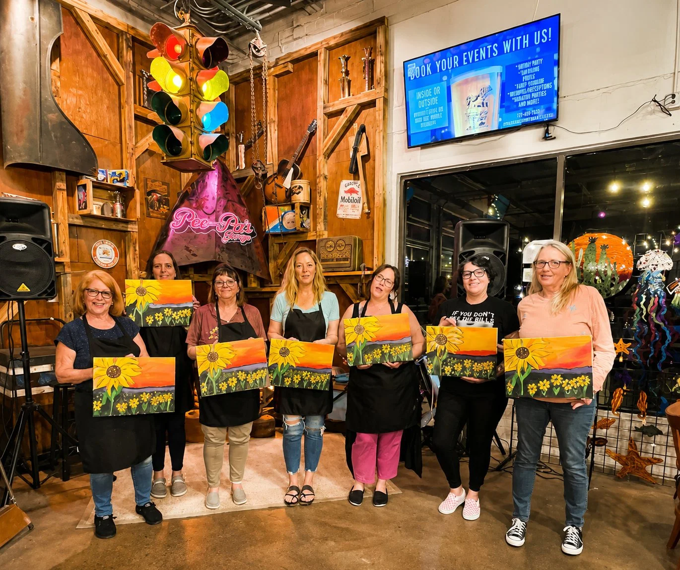 Six women standing in a row holding paintings of sunflowers and a sunset. They are inside a rustic venue with wooden walls, musical instruments, and a large screen displaying event booking information.