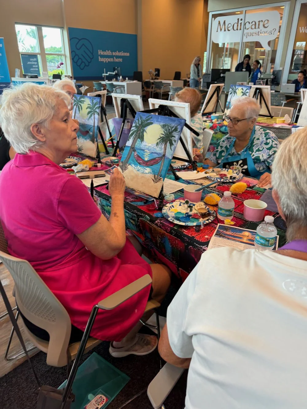 Older women painting tropical beach scenes on canvases during a group art activity in a community center or event space, with water bottles and painting supplies on the table.
