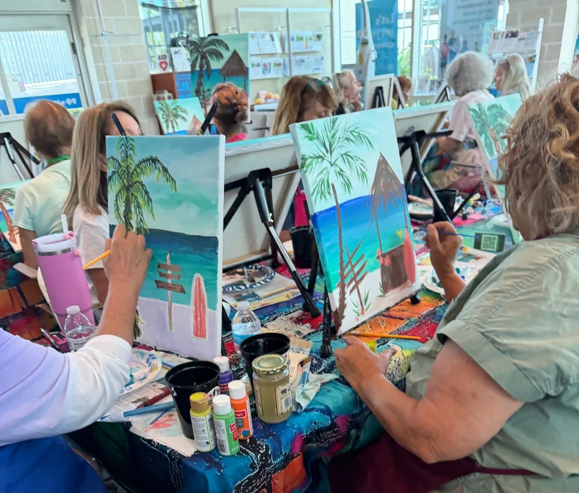 Group of older adults painting tropical beach scenes with palm trees and huts on canvas at an indoor art class.