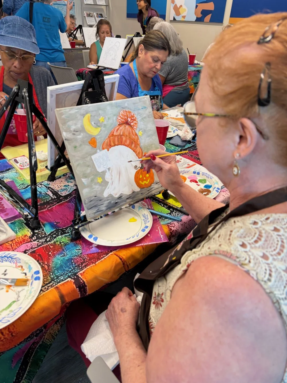A woman with glasses and red hair painting a Halloween-themed picture featuring a ghost, a pumpkin, a moon, and stars at a group painting event.