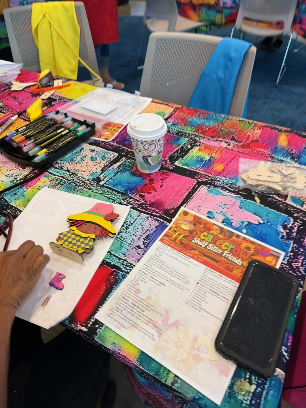 A colorful table setup with craft supplies, a coffee cup, a smartphone, and a paper titled 'Cearcrow Shelf Sitter Friends' at an arts and crafts event.