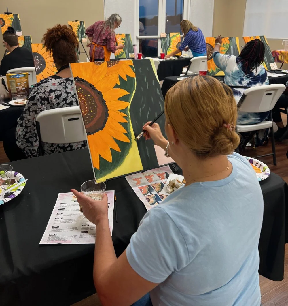Woman painting a sunflower on a canvas at an art class gathering.