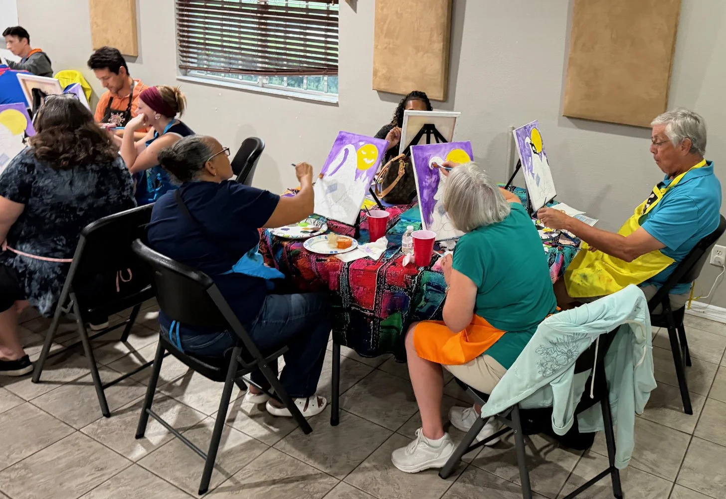 People participating in a group painting class, seated around a table with colorful tablecloth, painting canvases featuring a yellow sun and a purple background.