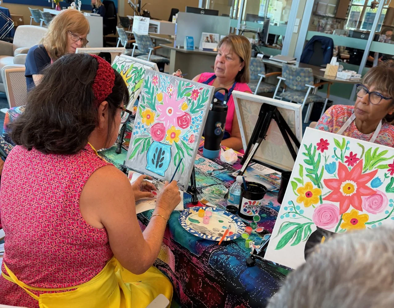 A group of women participating in a paint and sip class, painting colorful floral still life on canvases at a table in a bright, well-lit room.