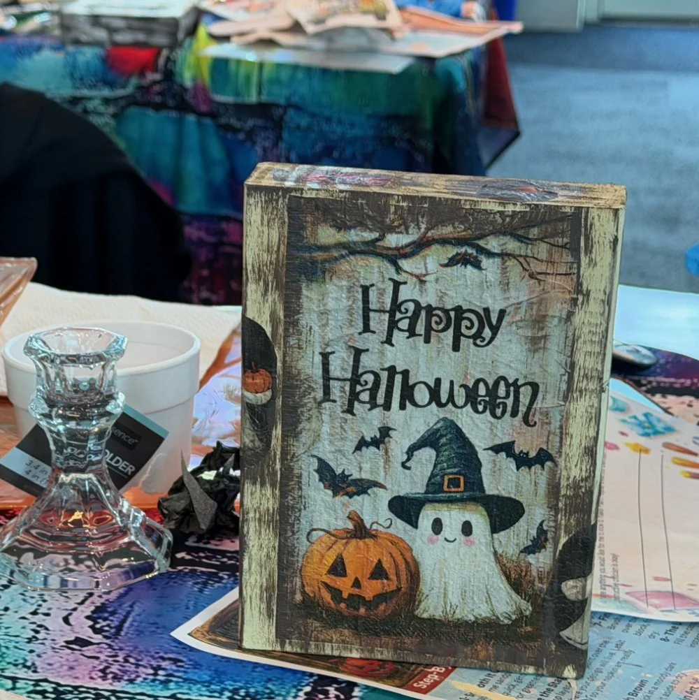 A Halloween greeting card featuring a ghost with a witch's hat, a carved pumpkin, and bats, with the words 'Happy Halloween' on it, placed on a table with various items.