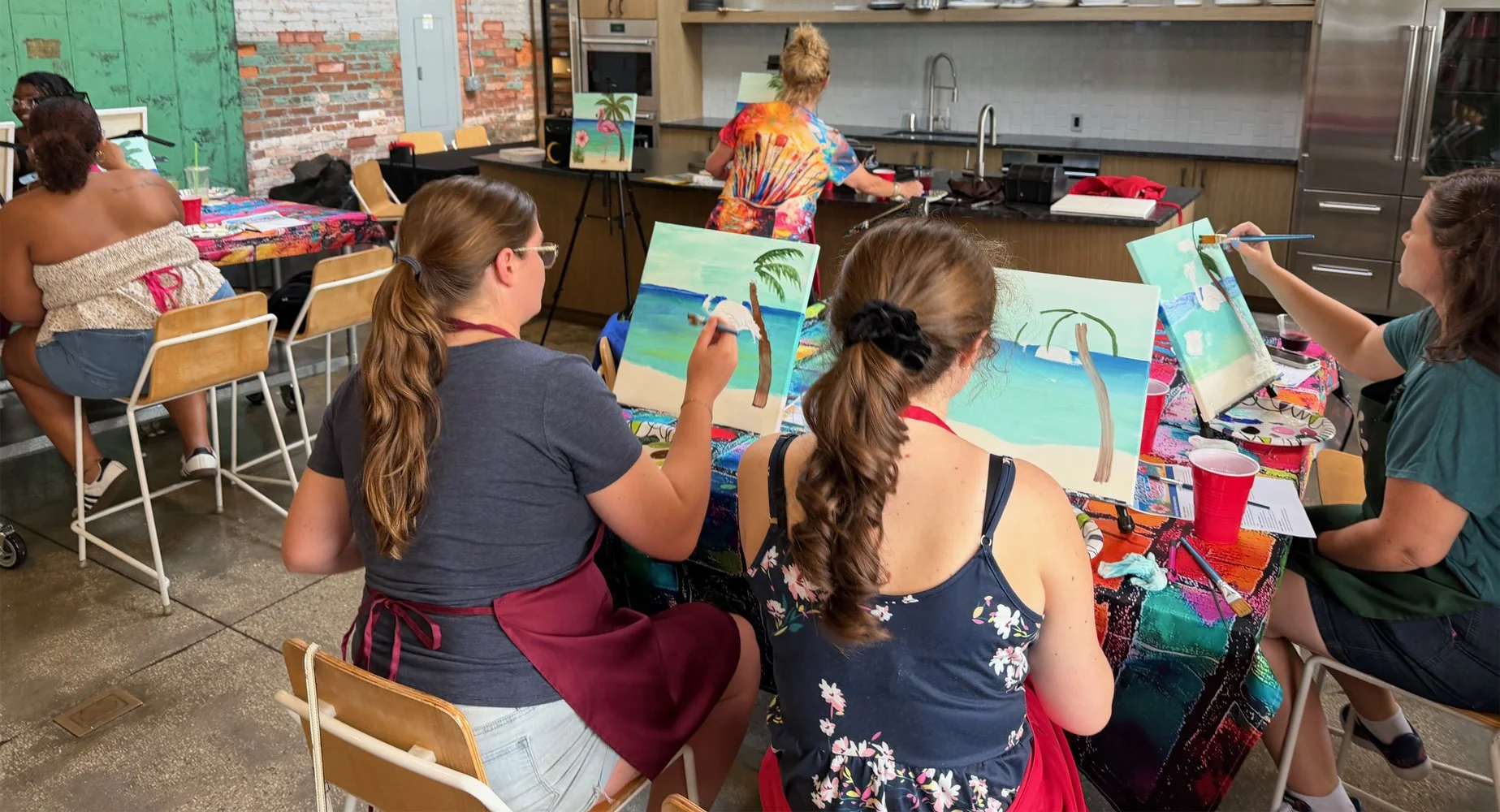 Group of women participating in a painting class, sitting at a colorful table with easels and canvases depicting tropical beach scenes, in an industrial-style room.