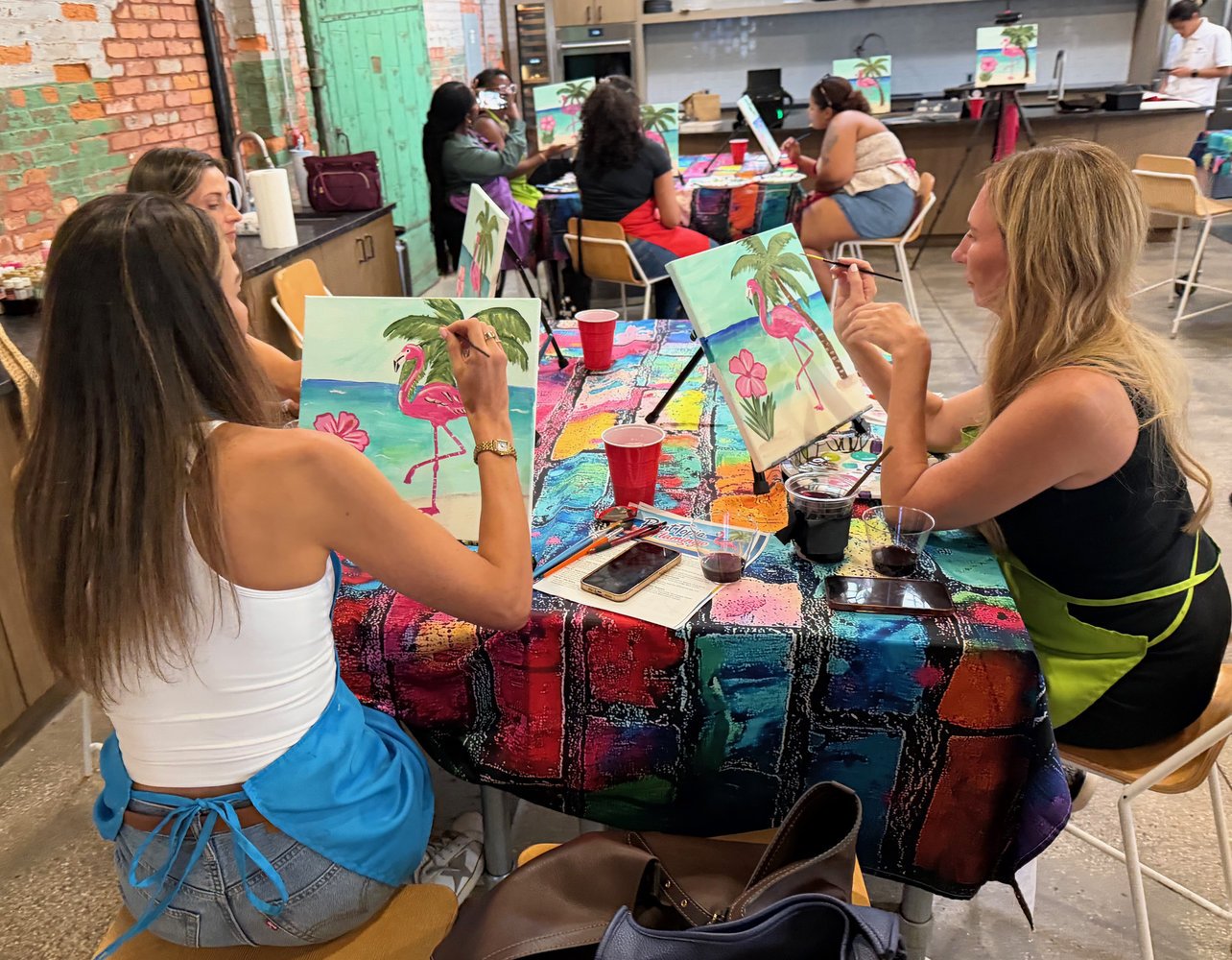 Women painting tropical-themed artwork with pink flamingos, palm trees, and flowers at a colorful table during a group art activity in a vibrant indoor studio.