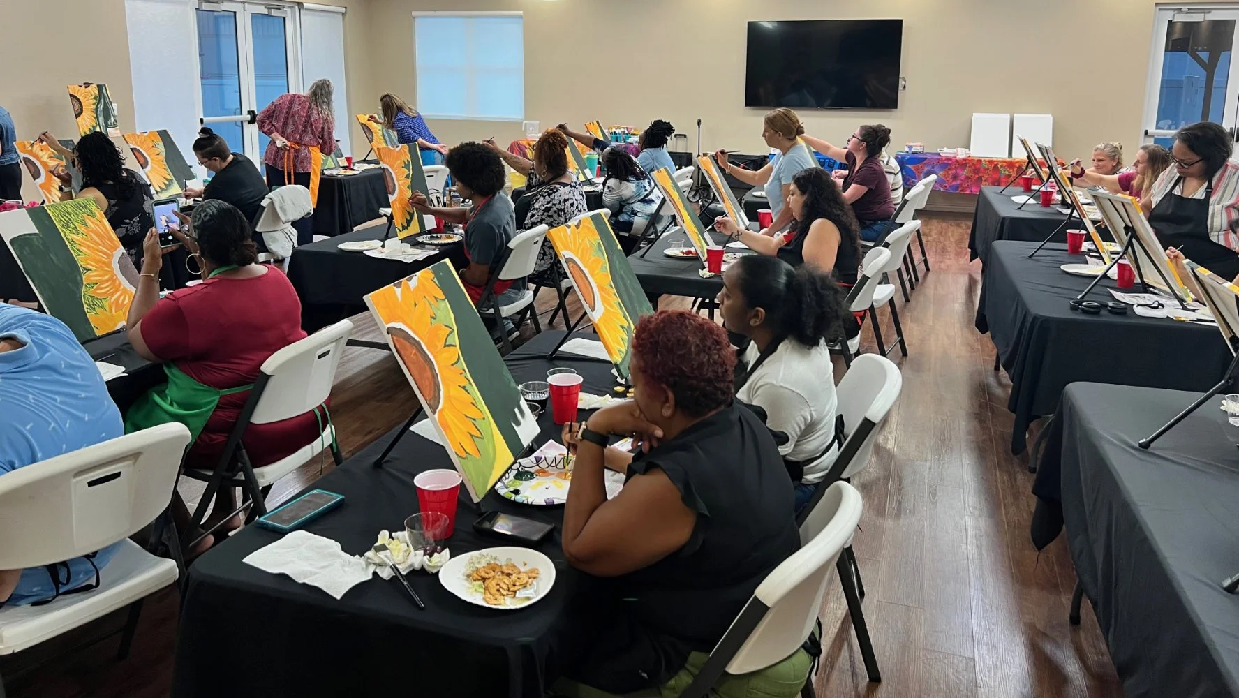 A group of people painting sunflower canvases in a classroom or art studio. They are seated at tables with painting supplies, and some are in the process of painting, while others are taking photos or observing. The room has large windows, a mounted 