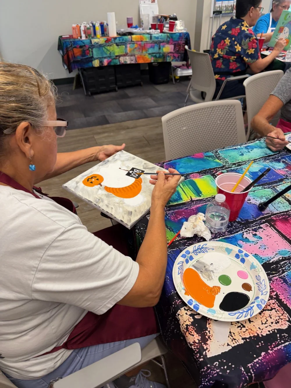 Woman painting a Halloween-themed pumpkin on a canvas at a paint-and-sip event. Art supplies and colorful paint palette on the table.